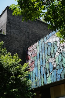 A modern building features an artistic facade with vibrant, abstract patterns in blue, green, and red. The structure is partially obscured by lush green trees, creating a contrast between the natural and man-made elements. Sunlight filters through the leaves, casting shadows on the dark brick wall.