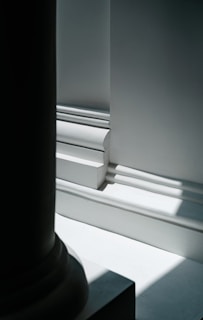 A close-up view of architectural details featuring strong light and shadow contrasts. The image shows intricate molding with clean lines and sharp edges, highlighting the smooth surfaces. The lighting creates a play of light and dark on the white and gray surfaces.
