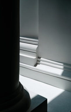 A close-up view of architectural details featuring strong light and shadow contrasts. The image shows intricate molding with clean lines and sharp edges, highlighting the smooth surfaces. The lighting creates a play of light and dark on the white and gray surfaces.