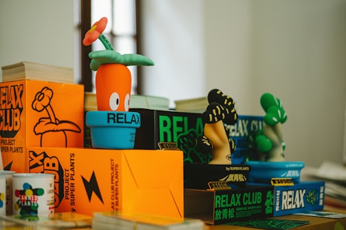 A group of colorful, cartoonish plant figures placed among vibrant boxes and books. The figures resemble plants with exaggerated features, including one with an orange pot and another with a whimsical flower on its head. The surrounding boxes are marked with bold text, reading 'RELAX CLUB' and are mostly in orange and green shades.