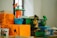 A group of colorful, cartoonish plant figures placed among vibrant boxes and books. The figures resemble plants with exaggerated features, including one with an orange pot and another with a whimsical flower on its head. The surrounding boxes are marked with bold text, reading 'RELAX CLUB' and are mostly in orange and green shades.