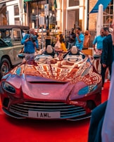 A sleek red sports car with a luxury design is parked on a red carpet. The setting includes several people observing and photographing the car, with some standing and others walking by. Buildings with large windows and awnings are visible in the background.