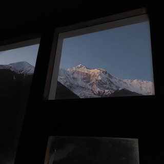 A snow-covered mountain peak is visible through a window, with the light of the sunrise casting a warm glow on the upper portions. The sky is clear and blue, providing a serene backdrop.