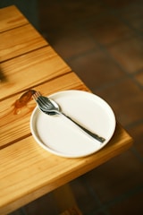 A wooden table with a small, round white plate resting on it. A stainless steel fork and spoon are placed in a crisscross pattern on the plate, with the spoon below the fork. The table has a visible darker knot in the wood grain and appears to be in a well-lit, indoor environment with a tiled floor in the background.