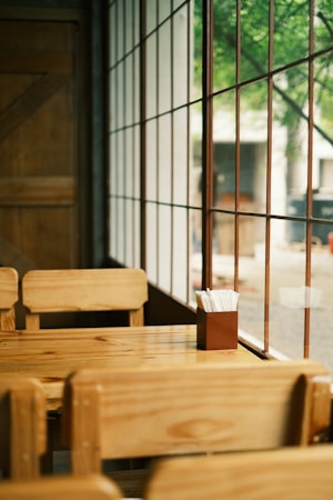 A wooden table and chairs are set in a cozy corner next to large windows with a grid pattern. Outside, trees are visible, creating a serene and natural view. The interior has a warm, inviting feel, with a focus on simple and natural materials.