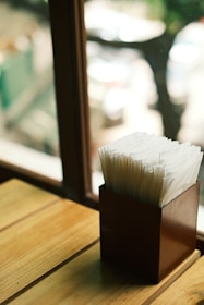 A wooden holder filled with neatly arranged white napkins is placed on a wooden surface. Natural light filters through a nearby window, casting soft shadows and creating a warm, serene atmosphere. The background appears slightly blurred, suggesting an outdoor setting beyond the window.