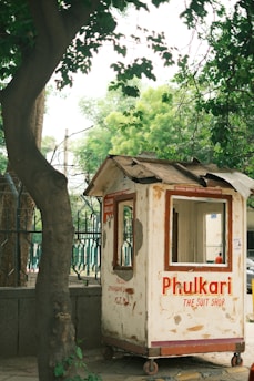 A small, weathered wooden kiosk painted white with red text stands on a sidewalk. The text reads 'Phulkari The Suit Shop' along with other faded writings. Surrounding the kiosk are green trees and a metal fence, creating a natural urban setting.