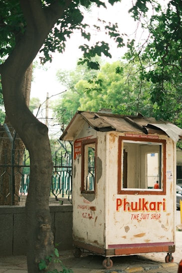 A small, weathered wooden kiosk painted white with red text stands on a sidewalk. The text reads 'Phulkari The Suit Shop' along with other faded writings. Surrounding the kiosk are green trees and a metal fence, creating a natural urban setting.