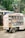 A person is standing on top of a delivery truck that is loaded with packaged beverages. The truck has compartments filled with various bottles, possibly soda or water, and is parked on a street next to a shop. The surrounding area has trees and shop signs, contributing to an urban setting.