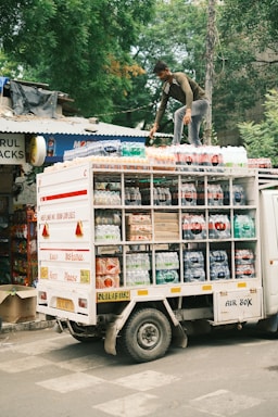A person is standing on top of a delivery truck that is loaded with packaged beverages. The truck has compartments filled with various bottles, possibly soda or water, and is parked on a street next to a shop. The surrounding area has trees and shop signs, contributing to an urban setting.