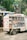 A person is standing on top of a delivery truck that is loaded with packaged beverages. The truck has compartments filled with various bottles, possibly soda or water, and is parked on a street next to a shop. The surrounding area has trees and shop signs, contributing to an urban setting.