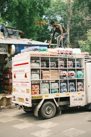 A person is standing on top of a delivery truck that is loaded with packaged beverages. The truck has compartments filled with various bottles, possibly soda or water, and is parked on a street next to a shop. The surrounding area has trees and shop signs, contributing to an urban setting.