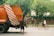 A person is placing branches into the back of an orange garbage truck on a street lined with trees. Another individual is riding a bicycle nearby. The surroundings are urban with a mix of greenery.
