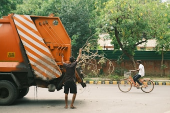 A person is placing branches into the back of an orange garbage truck on a street lined with trees. Another individual is riding a bicycle nearby. The surroundings are urban with a mix of greenery.