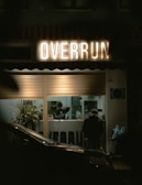 A dimly lit storefront with the illuminated sign 'OVERRUN' above a window showing a person inside, presumably a worker. There are green plants near the window and a parked car partially visible in the foreground. Another person is seated inside, facing away.