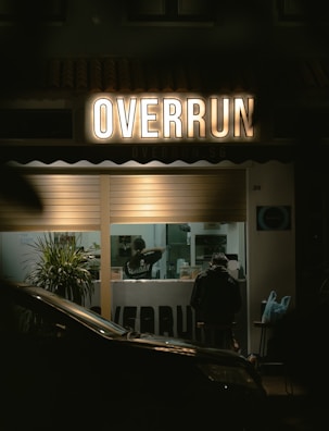 A dimly lit storefront with the illuminated sign 'OVERRUN' above a window showing a person inside, presumably a worker. There are green plants near the window and a parked car partially visible in the foreground. Another person is seated inside, facing away.