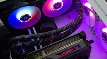 A close-up of a computer's internal components, including a graphics card labeled 'Gigabyte' and RGB cooling fans emitting vibrant colors. Cables are neatly arranged, and the lighting creates a modern, tech-savvy atmosphere.