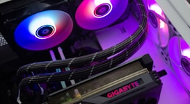 A close-up of a computer's internal components, including a graphics card labeled 'Gigabyte' and RGB cooling fans emitting vibrant colors. Cables are neatly arranged, and the lighting creates a modern, tech-savvy atmosphere.