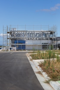 A modern building under construction surrounded by scaffolding. The site is fenced off, and there is a paved road leading up to the structure. Patches of grass and weeds grow on the sidewalk. The sky is clear and blue, suggesting a sunny day.