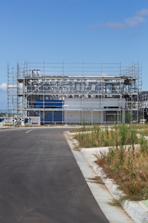A modern building under construction surrounded by scaffolding. The site is fenced off, and there is a paved road leading up to the structure. Patches of grass and weeds grow on the sidewalk. The sky is clear and blue, suggesting a sunny day.