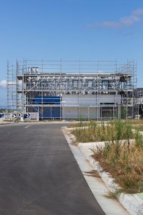 A modern building under construction surrounded by scaffolding. The site is fenced off, and there is a paved road leading up to the structure. Patches of grass and weeds grow on the sidewalk. The sky is clear and blue, suggesting a sunny day.