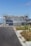 A modern building under construction surrounded by scaffolding. The site is fenced off, and there is a paved road leading up to the structure. Patches of grass and weeds grow on the sidewalk. The sky is clear and blue, suggesting a sunny day.