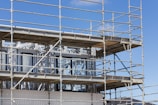 A building under construction is surrounded by scaffolding. The structure appears to have unfinished metal frames and partially installed windows. The blue sky serves as the backdrop.