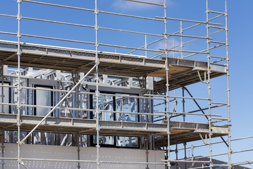 A building under construction is surrounded by scaffolding. The structure appears to have unfinished metal frames and partially installed windows. The blue sky serves as the backdrop.