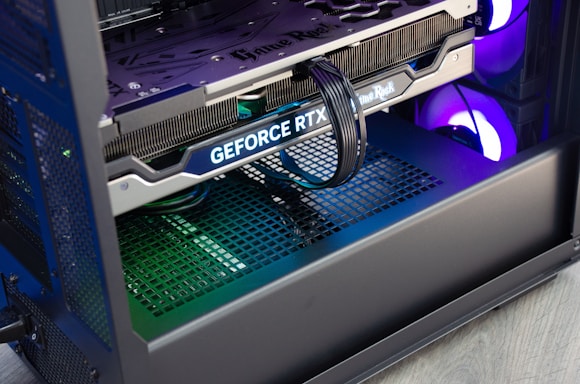 A close-up view of the interior of a computer casing showcasing a high-end graphics card with 'GeForce RTX' branding. The graphics card is secured in its slot, and several power cables are attached. The background reveals cooling fans illuminated with RGB lighting.