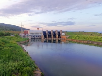 A landscape view features a large dam structure with spillways extending across a calm body of water. Surrounding the dam is lush green vegetation, and there is a clear blue sky with scattered clouds overhead. The area is peaceful and industrial, with utility poles visible.