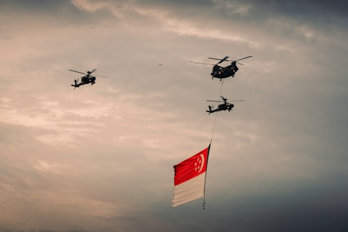 Three helicopters fly in formation with a large Singaporean flag suspended beneath the central helicopter against a cloudy sky.