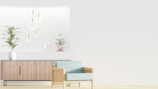 A minimalist interior design featuring a wooden sideboard with sleek lines and a potted plant on top. Modern pendant lights with circular designs hang from the ceiling, and decorative items are arranged on a recessed shelf. A light blue armchair with a wooden frame is positioned in the foreground, enhancing the contemporary aesthetic.