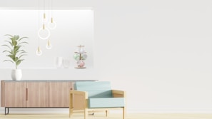 A minimalist interior design featuring a wooden sideboard with sleek lines and a potted plant on top. Modern pendant lights with circular designs hang from the ceiling, and decorative items are arranged on a recessed shelf. A light blue armchair with a wooden frame is positioned in the foreground, enhancing the contemporary aesthetic.