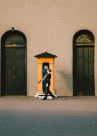 A uniformed guard is marching with a rifle over his shoulder. He is positioned in front of a brightly colored sentry box. The setting includes large doors and arched windows against a sandy-colored wall.