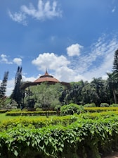 A lush, green garden with well-maintained hedges and trees surrounds a small pavilion with a traditional, ornate roof. The sky is bright blue and dotted with fluffy white clouds, creating a peaceful and serene landscape.