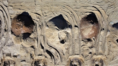 Aerial view of construction site soil with distinct tire tread patterns and uneven terrain. The surface shows a variety of textures and shades, primarily composed of light earth tones.