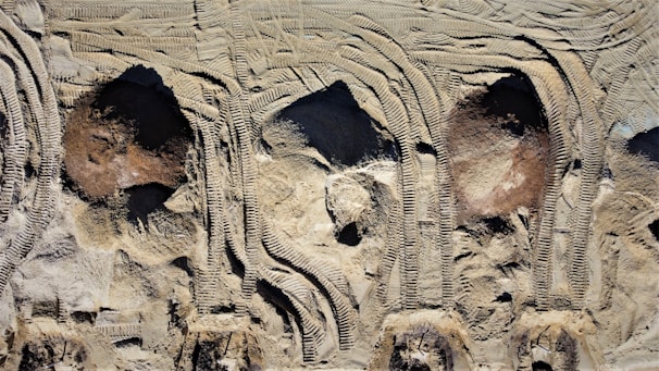 Aerial view of construction site soil with distinct tire tread patterns and uneven terrain. The surface shows a variety of textures and shades, primarily composed of light earth tones.