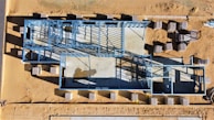 An aerial view of a construction site featuring a building foundation framed with blue steel beams. The site is surrounded by a sandy landscape, and several stacks of materials are scattered around.