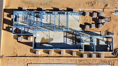 An aerial view of a construction site featuring a building foundation framed with blue steel beams. The site is surrounded by a sandy landscape, and several stacks of materials are scattered around.