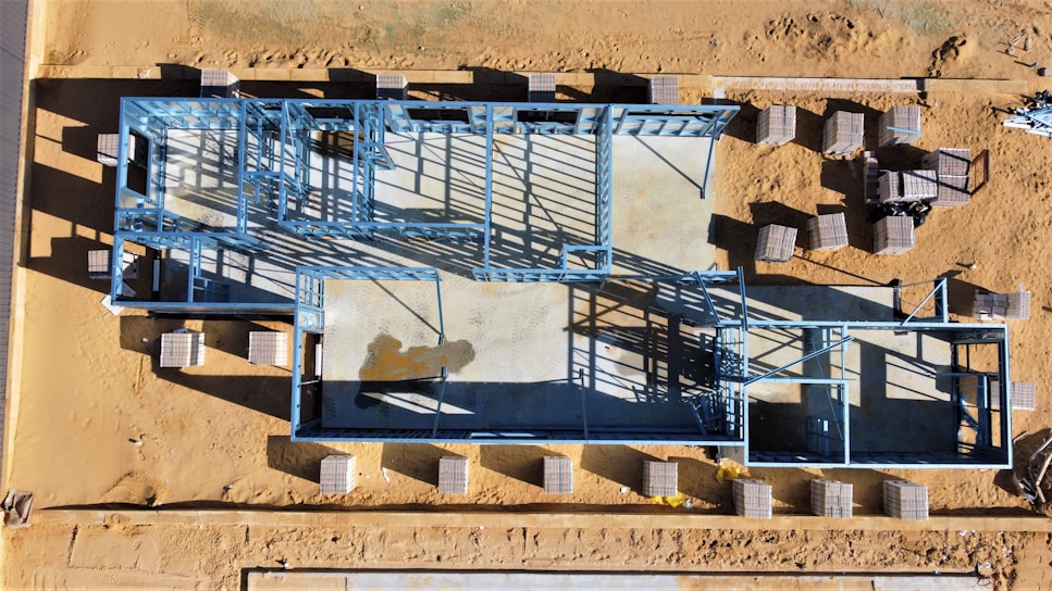 An aerial view of a construction site featuring a building foundation framed with blue steel beams. The site is surrounded by a sandy landscape, and several stacks of materials are scattered around.