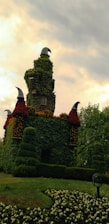 A whimsical tower covered in lush greenery and flowers stands against a cloudy sky. The tower features pointed, black cone-shaped structures atop it, creating a fairytale-like appearance. Surrounding the tower are neatly trimmed bushes and a flowerbed blooming with small white flowers.