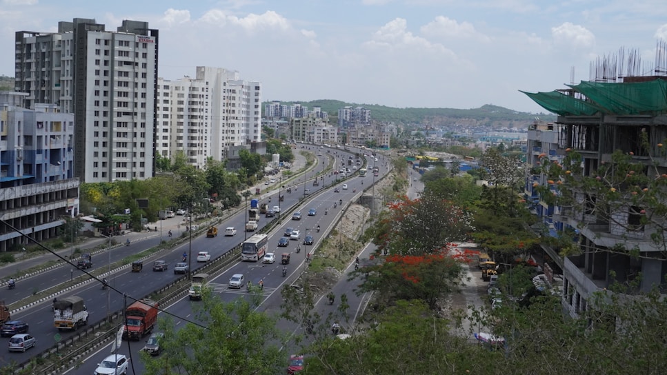 A busy urban highway is flanked by tall residential buildings on one side and a partially constructed building on the other. Multiple vehicles, including cars, buses, and trucks, are traveling in both directions along the multi-lane road. The area is lush with trees, and the landscape extends to a hilly horizon under a partly cloudy sky.