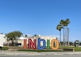 Colorful large letters spelling out 'INDIO' stand in an outdoor space with a backdrop of buildings and palm trees against a clear blue sky. A mix of greenery and street elements like a bench and a sidewalk are also visible.