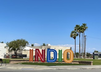 Colorful large letters spelling out 'INDIO' stand in an outdoor space with a backdrop of buildings and palm trees against a clear blue sky. A mix of greenery and street elements like a bench and a sidewalk are also visible.