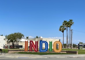 Colorful large letters spelling out 'INDIO' stand in an outdoor space with a backdrop of buildings and palm trees against a clear blue sky. A mix of greenery and street elements like a bench and a sidewalk are also visible.