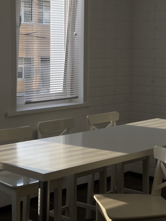 A modern dining area with a long white table and matching chairs is situated near a window with Venetian blinds. Soft sunlight filters through the blinds, casting striped shadows across the table and chairs. The room features white tiled walls and is minimally decorated.