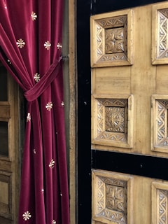 A rich red velvet curtain with embroidered floral patterns is elegantly draped beside a detailed wooden door featuring intricate geometric carvings. The door's natural wood tones are complemented by black accents framing the carved panels.