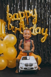 A young child is smiling while sitting on a small ride-on toy car. The background features gold balloons spelling 'happy birthday' and additional gold balloons on the side.