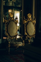 Two ornate, antique chairs with intricate gold detailing and upholstered seats are placed side by side in a dimly lit room with reflective surfaces and rich textures.