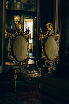 Two ornate, antique chairs with intricate gold detailing and upholstered seats are placed side by side in a dimly lit room with reflective surfaces and rich textures.